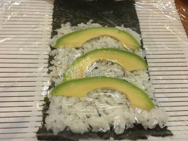 寿司帘上依次铺上海苔、米饭和片状鳄梨，然后上面铺一层保鲜膜，然后将海苔面翻面朝上；