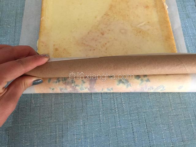 把擀面杖放在油纸的背面，用擀面杖推动着油纸把蛋糕卷起来。